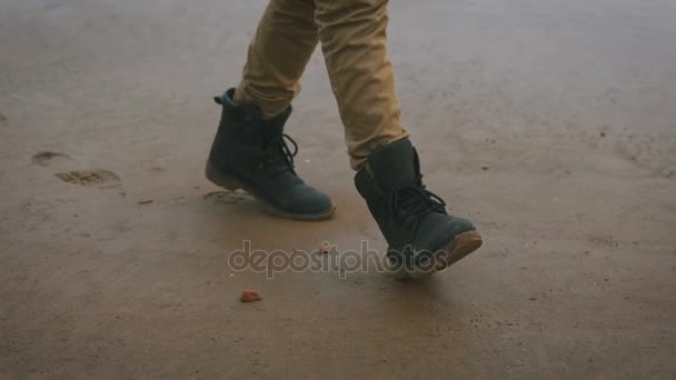 Zbliżenie na nogach młodego człowieka w gorczycy spodnie i czarne stylowe buty spaceru sam na plaży. Zwiedzanie chłopiec w ciężkich butów, pozostawiając ślady stóp na piasku. Piasek na plaży jest mokre i ciemny. — Wideo stockowe