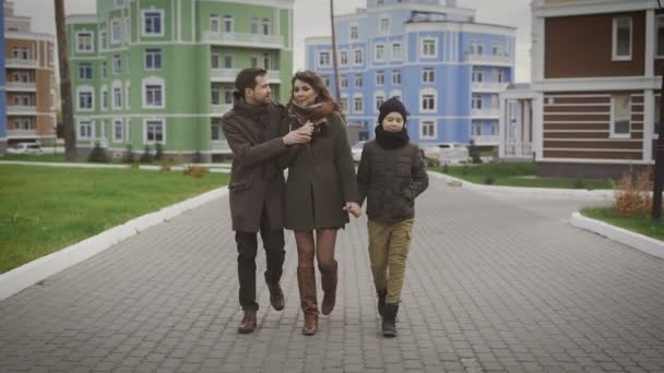 Tată fericit, mamă şi fiu făcând o plimbare împreună. Familia în haine calde merge împreună pe stradă cu clădirile de diferite culori în spatele lor. . — Videoclip de stoc