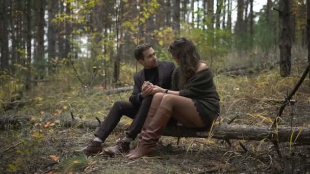 Close-up de casal feliz ter um encontro na floresta. Homem e mulher sorridentes estão sentados no tronco entre as árvores. Homem barbudo em camisola de pescoço de rolo preto está olhando amorosamente para sua mulher — Vídeo de Stock