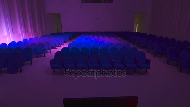 Modern concert hall with a lot of seats. Room off the main light and only spotlight with colored lights illuminate it. amera moves away from the seat in the hall — Stock Video