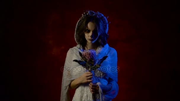 Close up of a ghost of young girl standing and spinning flower in her hands and moving her arm inviting to follow her. Phantom of a mystical woman having creative Halloween skull make-up. Dead girl is — Stock Video
