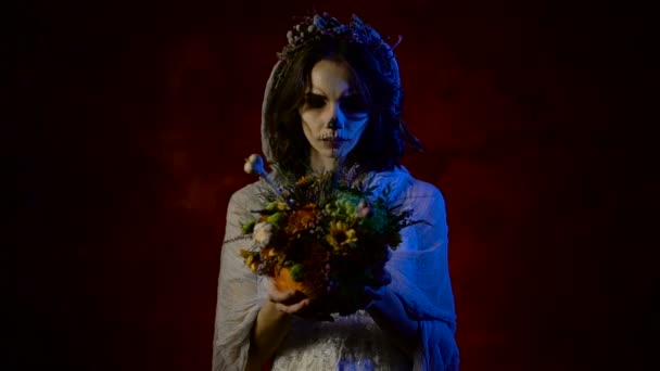 Close up of a ghost of a girl with bunch of flowers in her hands and looking at it. Young beautiful woman with art skull make-up on her face is participating in Halloween party