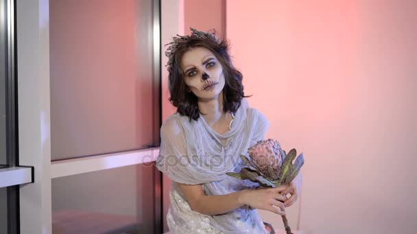 Закрытие молодой девушки с творческим пугающим гримом Хэллоуина. на вид мертвая невеста, сидящая на лавочке у окна с цветочком в руках и смотрящая в камеру . — стоковое видео