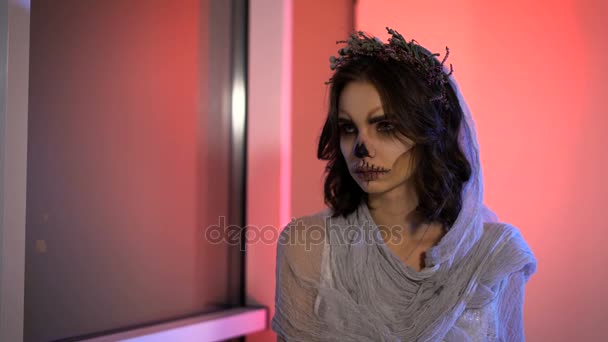 Gros plan du visage une jeune fille avec masque mort crâne art sur elle. mariée créative effrayant et effrayant Halloween faire est assis en regardant la fenêtre. le banc le fond rose . — Video