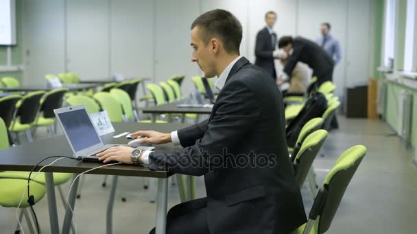 Hombre joven en traje de negocios con reloj de pulsera caro está trabajando con el ordenador portátil sentado en la silla verde detrás de escritorio negro en el aula con el grupo de personas en el fondo — Vídeos de Stock