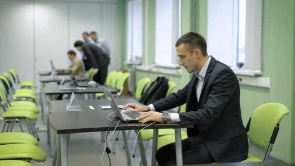 Młody człowiek w garniturze, siedzący na widowni i działa na laptopie. Przygotowanie uczestnika seminarium biznesowe przed wykład. Ludzie w tle wyglądają jak ich kolega z pracy — Wideo stockowe