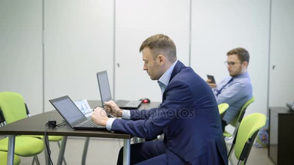 Jóvenes hombres de negocios en trajes de salón con computadoras portátiles están sentados en una silla verde detrás de un escritorio negro en el aula. Un hombre de anteojos con barba en el fondo está escribiendo y enviando mensajes por teléfono inteligente . — Vídeos de Stock