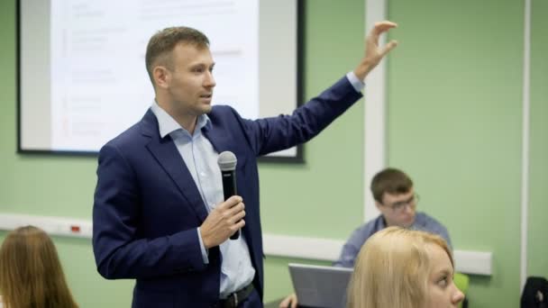 El profesor da conferencias sobre economía en la universidad. Con micrófono en la mano y con la ayuda de gestos, transmite a los estudiantes la información importante . — Vídeos de Stock