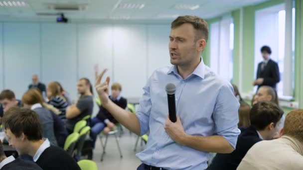 Profesionální trenér v modré košili, mluvit do mikrofonu a rukama na workshop pro budoucí top manažery obchodní společnosti. Publikum sedí v konferenčním sále v tabulce — Stock video