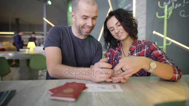 Casal jovem sentado em uma mesa e olhando para a tela do smartphone, clicando sobre ele com os dedos. Homem e mulher revisando suas fotos no gadget ao rir e comentar ativamente sobre eles . — Vídeo de Stock