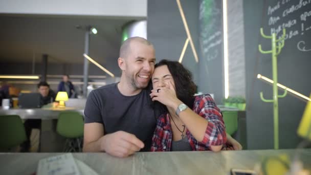 Jovem menina de cabelos pretos vestida com uma camisa vermelha e um homem barbudo em uma camisa cinza, sentados perto uns dos outros no café à mesa, rindo, brincando, segurando a mão uns dos outros e desfrutando — Vídeo de Stock