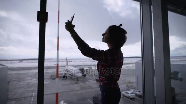 空港でショートヘアの美しいブルネットの少女。彼女は幸せです。彼女は休暇と旅行する計画を行っていたので、機嫌が良いです。Selfie は、空港のパノラマ ビューの窓に立っています。 — ストック動画