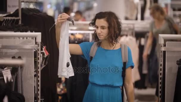 Schönes Mädchen in hellblauem Kleid geht einkaufen und hat die Wahl zwischen weißem T-Shirt oder grauem Hemd. junge Konsumentin trifft Entscheidung über den Einkauf im Duty Free Shop. — Stockvideo