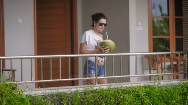 Odasından dışarı geliyor. Veranda için devam ediyor. Bali hava harika. O bir veranda için açık manzaralarına sahiptir. Bir Hindistan cevizi kokteyl elinde shes.