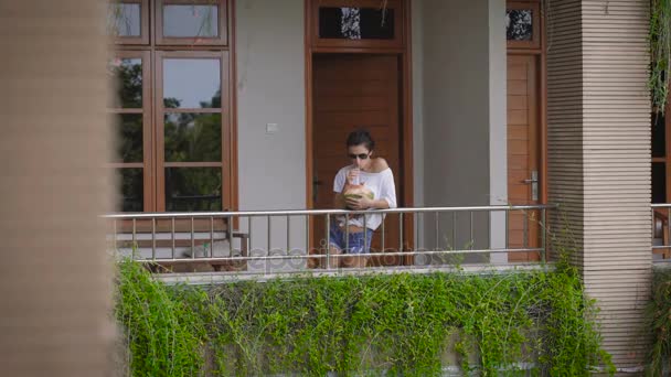 La ragazza con gli occhiali da sole è uscita sul balcone della sua camera per bere un cocktail esotico dal cocco. Turista in vacanza in un accogliente hotel . — Video Stock