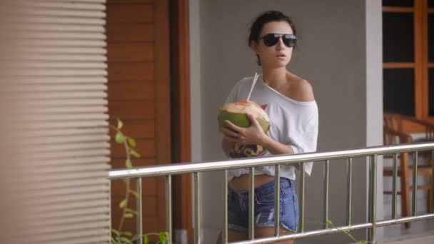Jong meisje in zonnebril in de ochtend permanent met de kokos en het drinken van het SAP door de rietjes. Stijlvolle vrouwelijke reiziger proeven van vers Balinese fruit op het terras van haar tropische huis. — Stockvideo