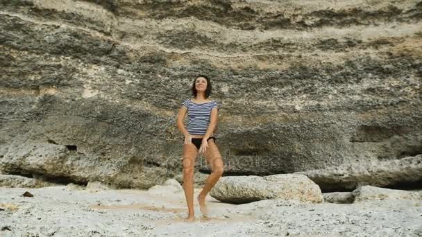 Stranden på ön Bali. Flickan inte lång brunett i en baddräkt. Shes på gott humör. Skämt, skratt, hoppar och dansar poserar för kameran. Bakgrund av en enorm stenmur. — Stockvideo