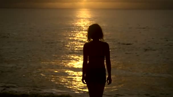 Струнка дівчина повільно ходить в океан на заході сонця, щоб охолодитися. Силует молодої жінки на тлі сонячних роздумів у воді, які піднімають руки вгору . — стокове відео