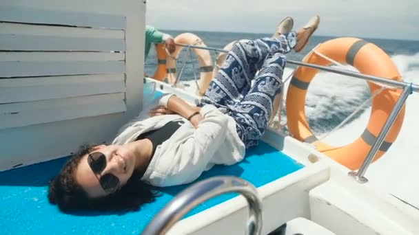 Glad flicka är att ha kryssning ombord på en yacht i det öppna havet. Leende kvinna liggande på däcket lite fartyg i havet med benen i luften mellan två orange liv-boj ringar. — Stockvideo
