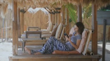 Güzel bir tembel bir tropikal resort Bali üzerinde yatan kız giyinmiş. Genç kadın otelde dinlenirken tablet ve akıllı telefon kullanır.