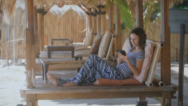 Die junge Frau entspannt sich an heißen Sommertagen auf Liegestühlen unter dem Zelt und bedient ihr Tablet. attraktive Brünette genießt ihren exotischen Urlaub und ruht sich allein mit ihrem Taschenbuch im Festzelt aus. — Stockvideo