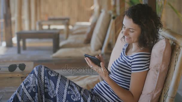 Schattig gelooid meisje in een striped T-shirt en blauwe broek zittend op een stoel buiten en het lezen van een verhaal over het boek van ellektronnoy met een glimlach — Stockvideo