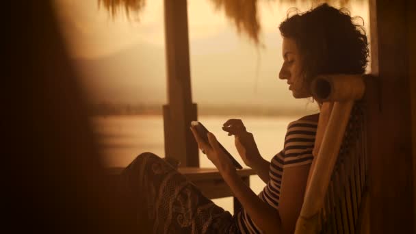 Zonsondergang en het strand op het eiland Bali. Het meisje in een gestreepte blouse en brede broeken in de avondzitting op een zachte stoel aan het strand. Hij speelt op de Tablet PC. — Stockvideo