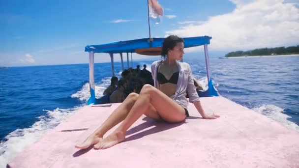 Primer plano de la atractiva joven posando en la cubierta de un pequeño yate en mar abierto. Joven modelo vestida con traje de baño negro está sentada a bordo y bañándose al sol disfrutando de su crucero . — Vídeo de stock