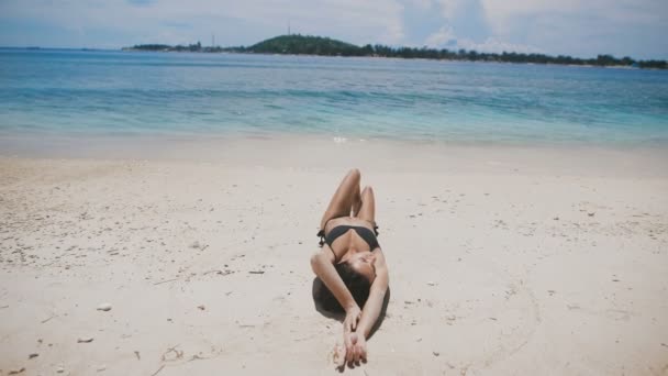 Сексуальная загорелая девушка с красивой грудью, лежащая на песке у океана и медленно опускает ногу, а затем рисует круг — стоковое видео