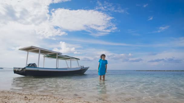 Piękna dziewczyna w niebieskiej sukience, chodzenie po wodzie w pobliżu zacumowane łodzie. Szczupła brunetka spacery po blisko piaszczystej plaży. Młoda kobieta relaksujący na tropical resort. — Wideo stockowe