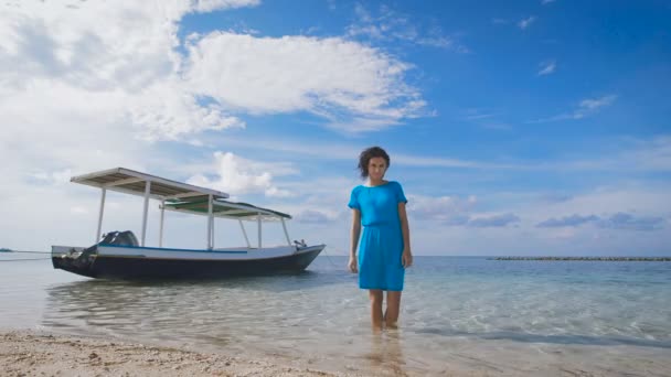 Une jolie brune vêtue de turquoise se tient dans la mer avec un bateau balinais traditionnel en arrière-plan. Fit belle fille se détendre dans l'océan avec un paysage marin à couper le souffle derrière . — Video
