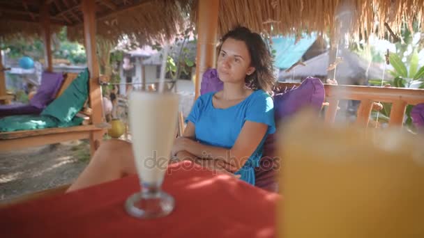 Junge schöne Brünette genießt ihren Urlaub in der Bar der tropischen Insel am Tisch sitzend. Reisende verbringt ihren Urlaub entspannt im Café unterm Strohdach und probiert Cocktails. — Stockvideo