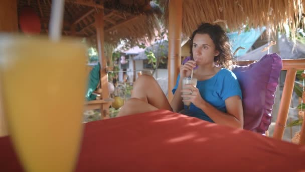 Brunetka z krótkimi włosami, siedząc na ławce w kawiarni na wyspie. Ona jest bardzo skupiony i wygląda na odległość, opierając się na poduszce picia koktajl. Na stole jest inny ananas koktajl — Wideo stockowe