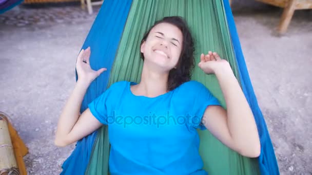 Χαρούμενη κοπέλα ξαπλωμένη στην αιώρα στην τροπική παραλία, κρύβοντας το πρόσωπό της και ευτυχώς το γέλιο. Αξιαγάπητη γυναίκα στο μπλε φόρεμα χαμογελαστός και χαλαρώνοντας σε swing ιστού σε μια ithe μπαρ του Μπαλί. — Αρχείο Βίντεο