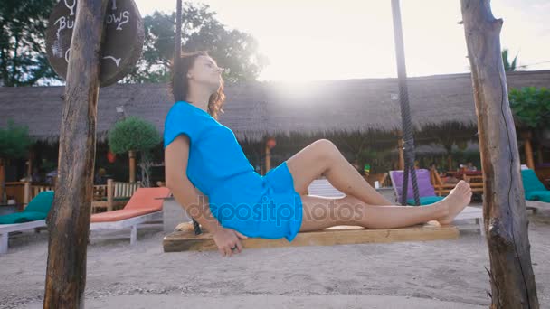 Attraktive Brünette in türkisfarbener Kleidung, die auf Schaukeln am balinesischen Strand mit tropischem Haus und Liegestühlen im Hintergrund spielt. junge Reisende vertreibt sich die Zeit auf faule Weise. — Stockvideo