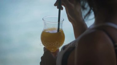 Kız bir kamışla içme turuncu kokteyl ve karıştırma içki. Sıvı kişi tarafından kullanımı kapat. Şeffaf bardak taze meyve suyu.