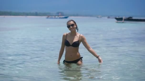 Hermosa morena con el pelo mojado de pie hasta la cintura en el agua y lleva en ella por las manos. Mujer joven en traje de baño negro y gafas de sol disfrutando de unas vacaciones en un resort tropical . — Vídeo de stock