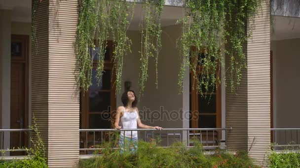 Hermosa viajera femenina está caminando en el balcón de su villa tropical y respirando aire fresco balinés. Joven morena está disfrutando de su viaje de pie en la terraza y mirando a su alrededor feliz . — Vídeos de Stock