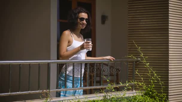 Mädchen mit Sonnenbrille kam auf den Balkon seines Zimmers, um ein Glas Wasser zu trinken. junge Frau in heller Hose und weißem Hemd schaut regelmäßig in die Ferne und lächelt. — Stockvideo