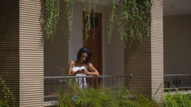 Piękne kobiece podróżnik stojąc na balkonie w swojej willi i jej smartfona. Młoda brunetka korzystających z jej podróży i czeka z przyjaciółmi na tarasie domu na wycieczkę po okolicy. — Wideo stockowe