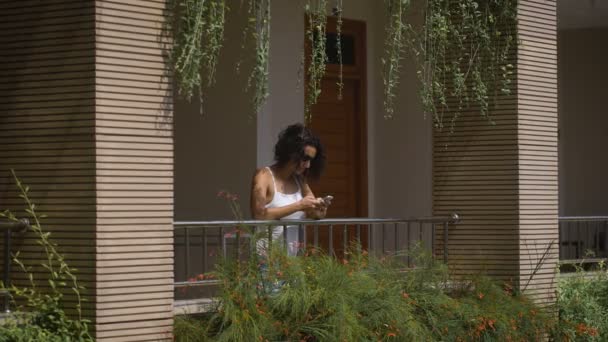 La fille aux cheveux noirs en lunettes de soleil debout sur le balcon du bâtiment, d'où pendent une plante, appuyé sur la balustrade et écrit un message à un ami au téléphone — Video