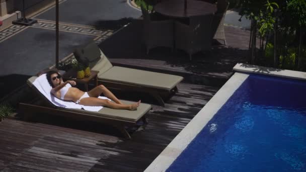 Na terytorium hotelu. Dziewczyna w białym kostiumie kąpielowym, leżąc na leżaku przy basenie. Rozluźnia i pobiera przyjemność z ich święta. Pogoda w Bali nie zawiedzie, ponieważ świeci słońce — Wideo stockowe
