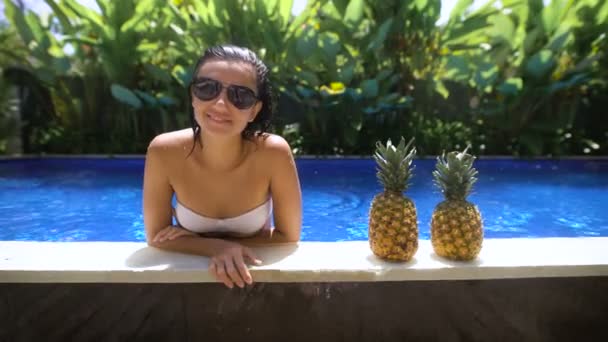 Красивая молодая девушка в жаркую погоду находится в бассейне и опирается на сцену, где есть два ананаса на фоне кустов — стоковое видео