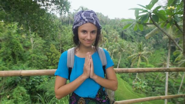 Тихая женщина с короткими темными волосами, носящая шарф на голове, голубую рубашку и красочный шарф, стоящая на деревянном мосту в тропическом лесу и прижимающая руки к груди, что означает благодарность — стоковое видео