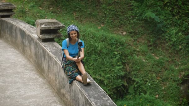 Une voyageuse souriante assise sur les balustrades du pont sur la terrasse et tenant un selfie-stick dans ses mains. Attrayant brunette essaie de faire selfie photo sur un fond de plantes tropicales . — Video