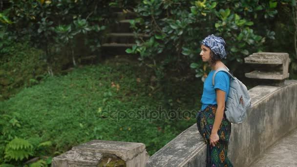 Mujer solitaria con cabello negro, velo en la cabeza, vestida con una camiseta azul y pañuelo atado alrededor de las piernas, de pie en el puente de piedra en medio de la selva verde y mirando pensativamente en — Vídeo de stock