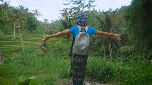 Russische Reiserin in lässiger blauer Kleidung steht inmitten endloser indonesischer Felder, hebt die Hände und genießt üppige tropische Vegetation und frische Luft.. — Stockvideo