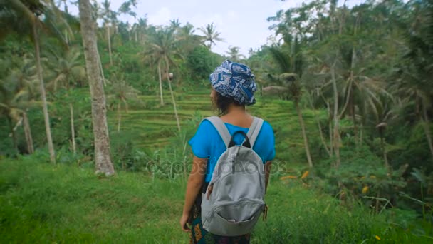 Braungebranntes Mädchen in blauem T-Shirt, Kopftuch und Rock mit Rautenabdrücken steht im Regenwald und blickt zurück in die Kamera, die eine graue Aktentasche trägt und sich unter den Palmen umsieht — Stockvideo
