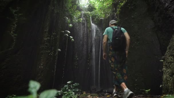 Jovem em um boné e uma mochila atingiu uma cachoeira incrivelmente bonita na selva. Turista desfrutando da beleza intocada da natureza . — Vídeo de Stock