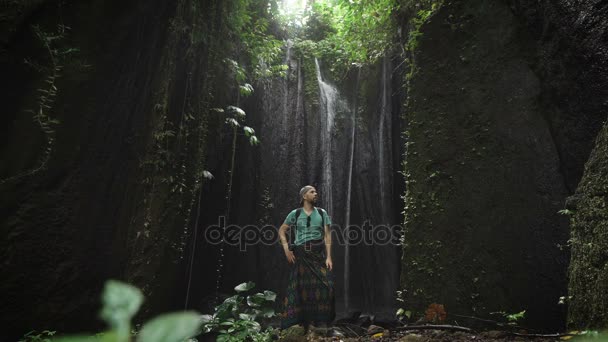 Elegante explorador do sexo masculino descansando em uma rocha na caverna perto de uma cachoeira atrás dele. Jovem de pé na baía dentro da montanha. Viajante relaxante dentro do jardim botânico tropical com cascata de resfriamento . — Vídeo de Stock